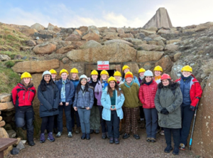 The field camp cohort group wearing colourful jackets and yellow hard hats, standing in front of the exit to Geevor Tin Mine